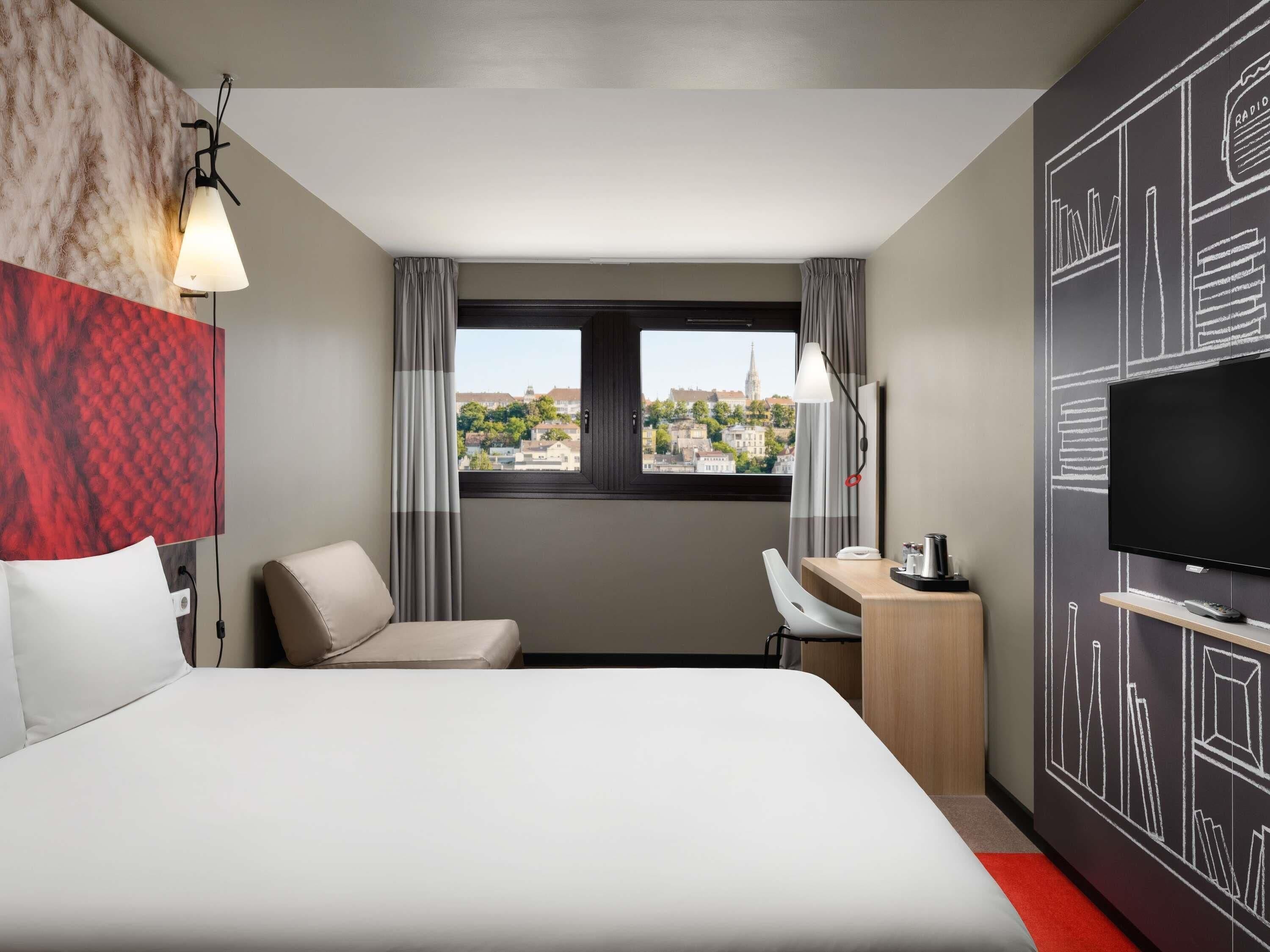 A hotel room with a bed, a desk, a chair, a television, and a window with a city view.