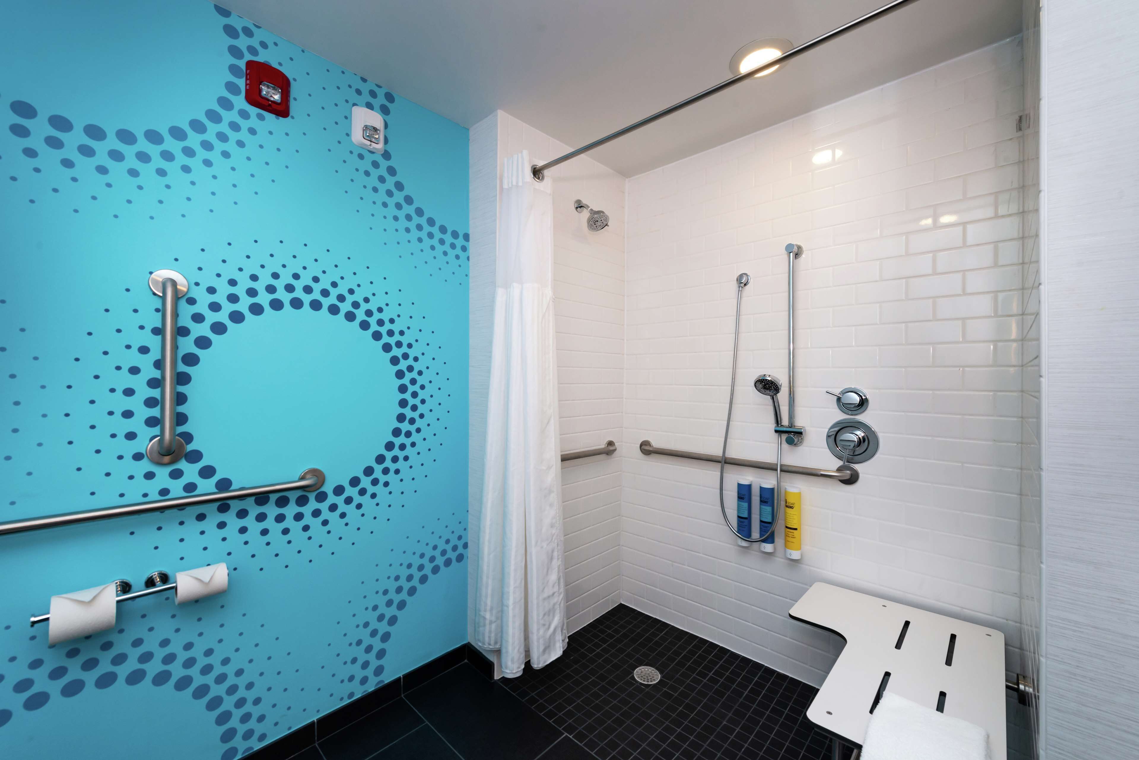 Um banheiro com parede azul, piso de azulejos brancos, chuveiro com barra de apoio e pia branca.