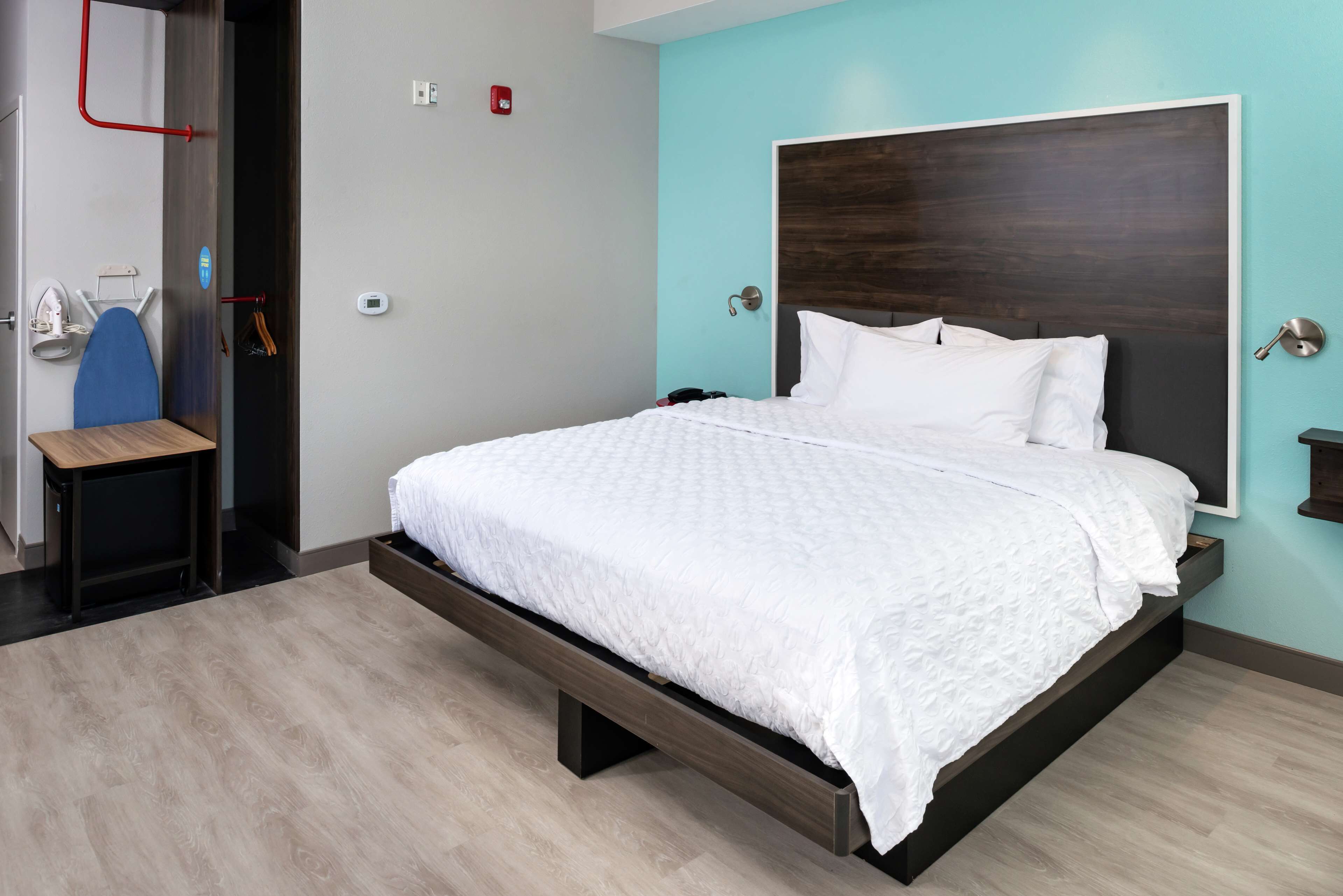 A modern bedroom with a large bed, a wooden nightstand, a desk with an ironing board, and a wall-mounted light.