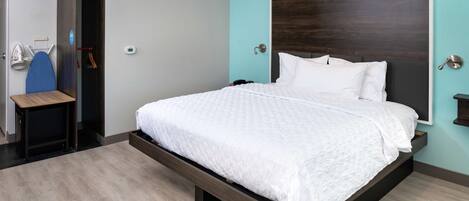 A modern bedroom with a large bed, a wooden nightstand, a desk with an ironing board, and a wall-mounted light.
