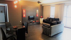 A modern living room with a flat-screen TV, a sofa, a dining table with chairs, and a large window with curtains. - PJ8 Service Suite Pool View (Petaling Jaya)