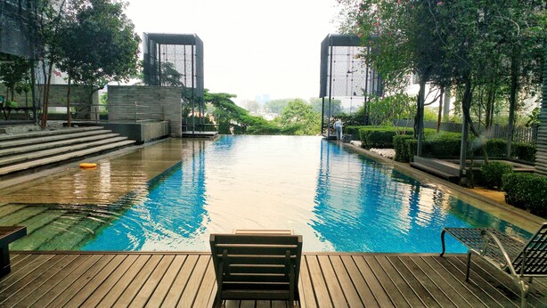A swimming pool with a wooden deck, steps leading into the water, and a view of a cityscape in the background. - PJ8 Service Suite Pool View (Petaling Jaya)