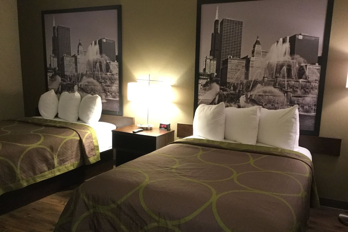 Habitación de hotel con dos camas, cada una con una colcha verde estampada, almohadas blancas y una mesita de noche con una lámpara entre ellas.