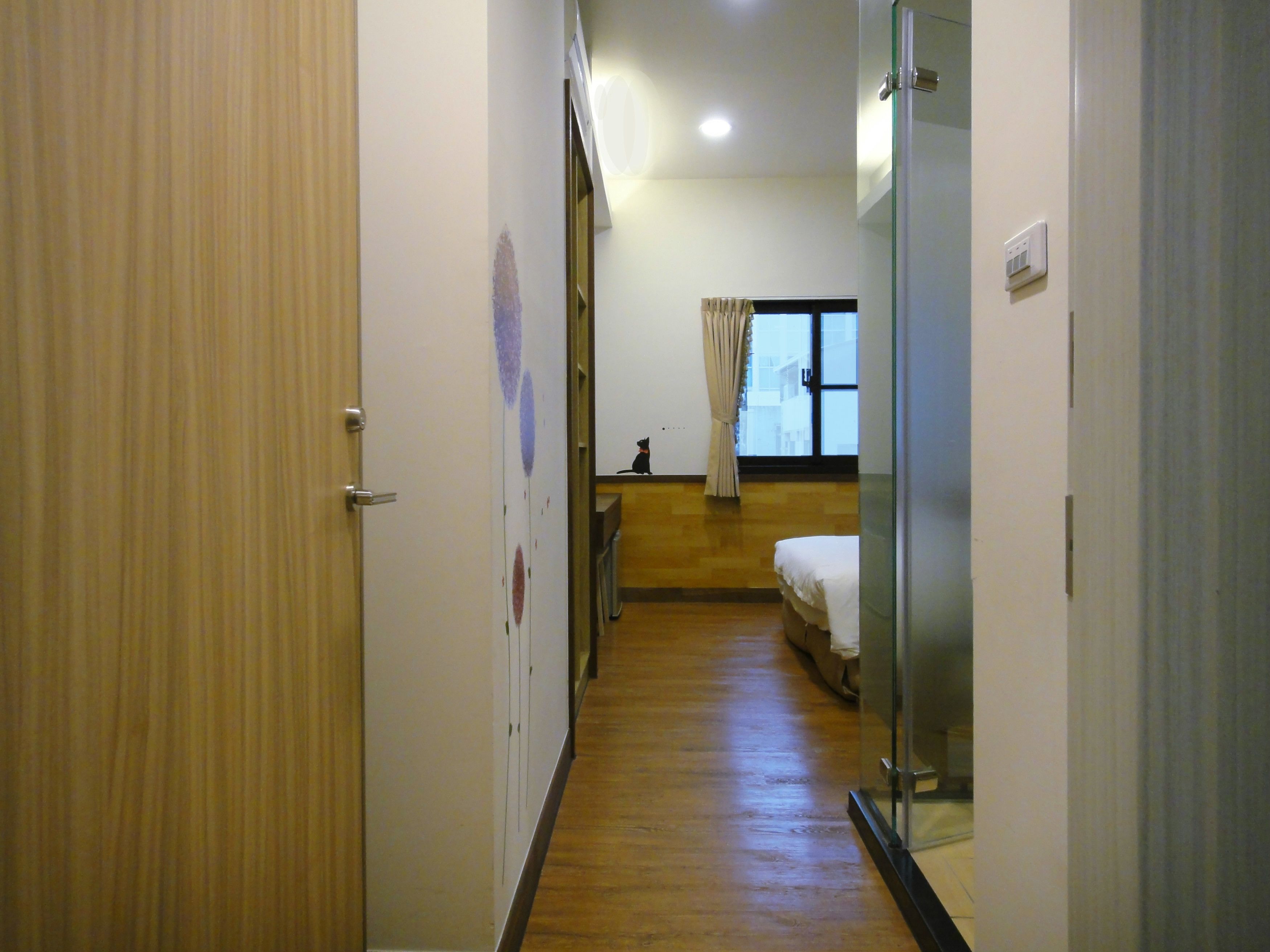 A narrow hallway with wooden flooring leading to a bedroom with a bed and a window.