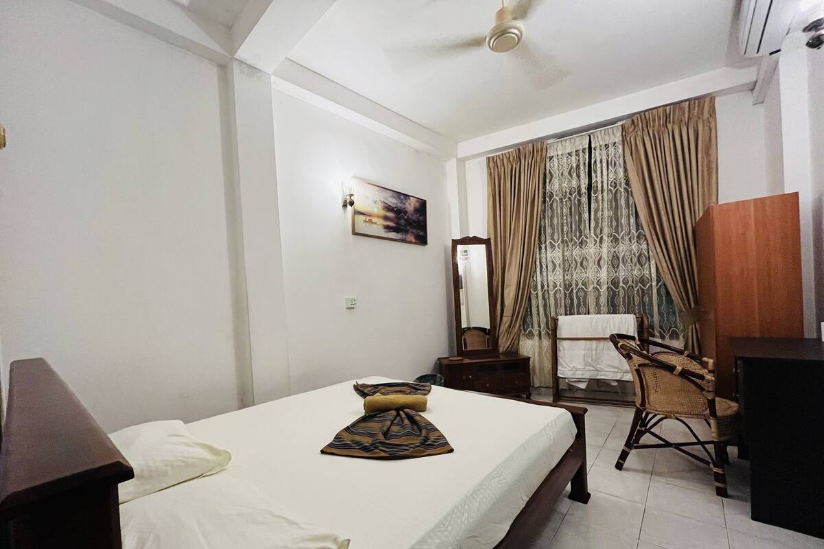 A bedroom with a bed, a chair, a desk, a mirror, and a window with curtains.