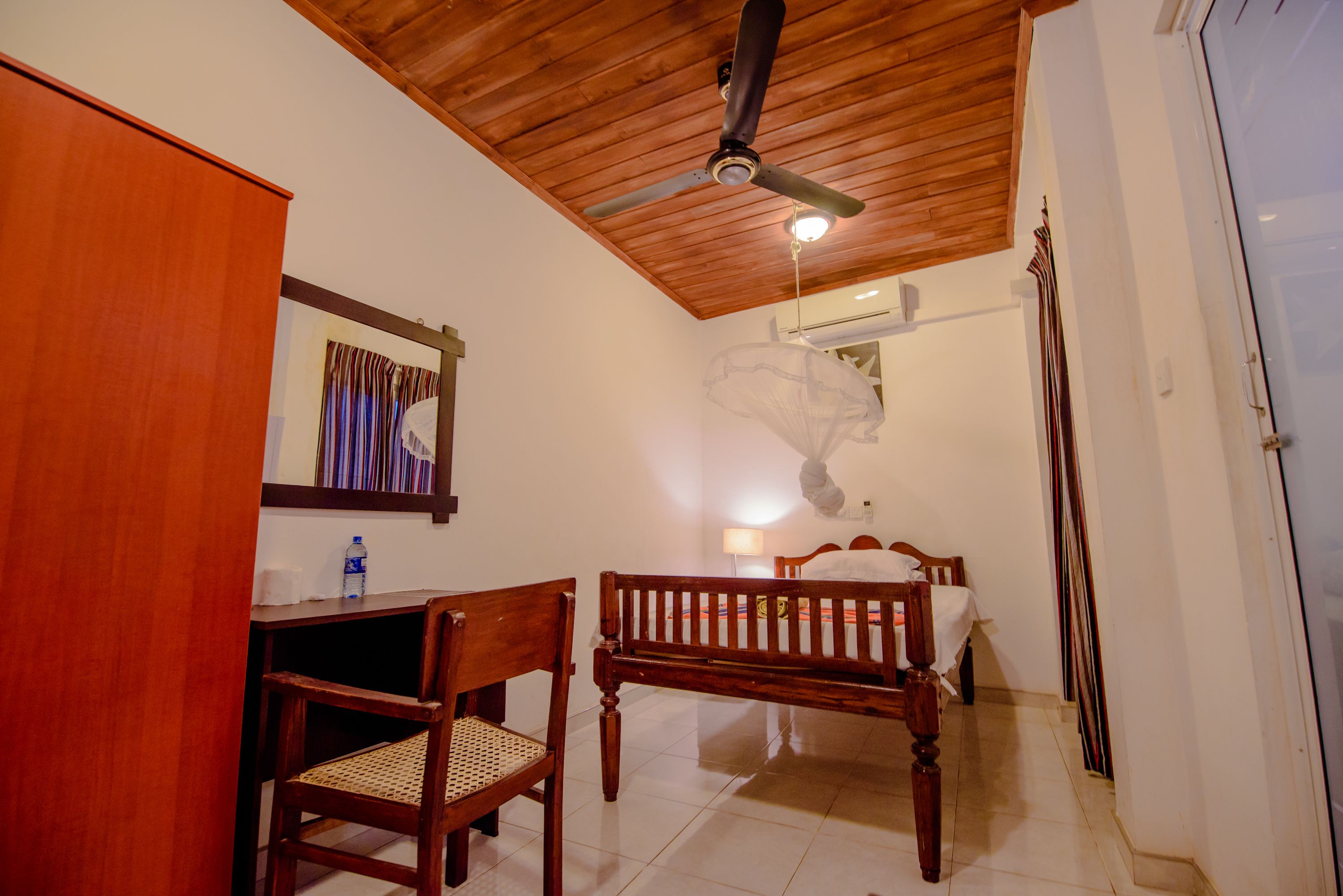 Una habitación con techo de madera, una cama con cubrecama blanco, un escritorio de madera con una silla y una botella de agua sobre el escritorio.