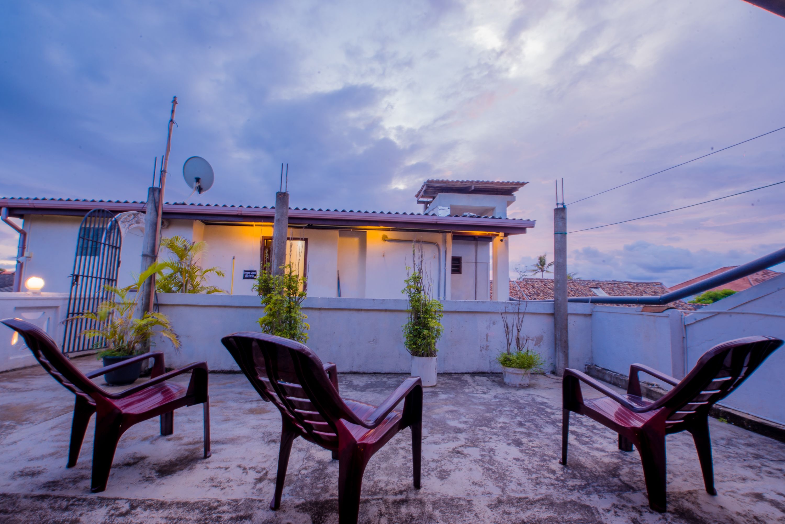 A rooftop terrace with three plastic chairs, potted plants, and a building with a satellite dish.