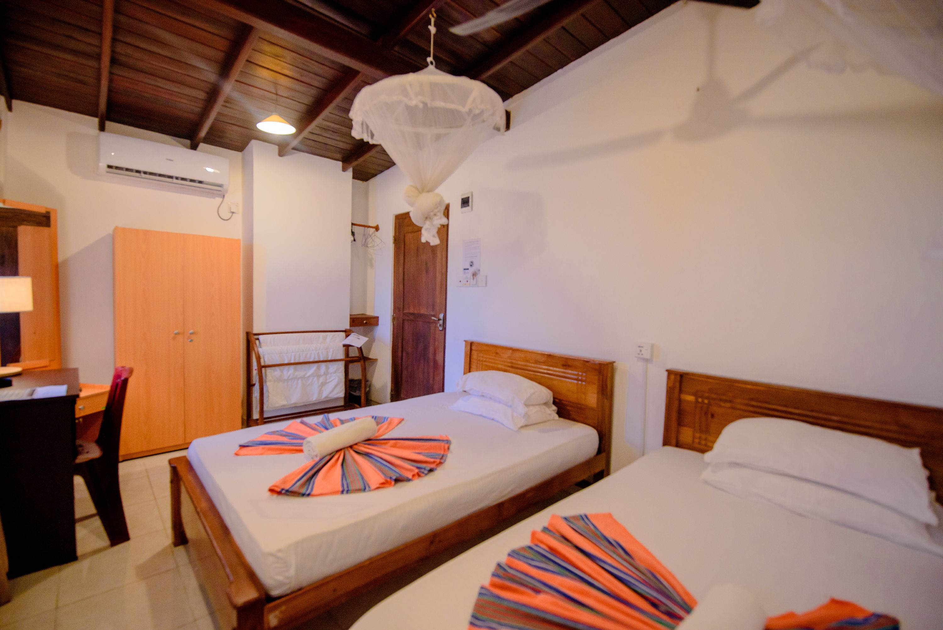 A hotel room with two beds, a wooden desk, a chair, a wardrobe, and a ceiling fan.