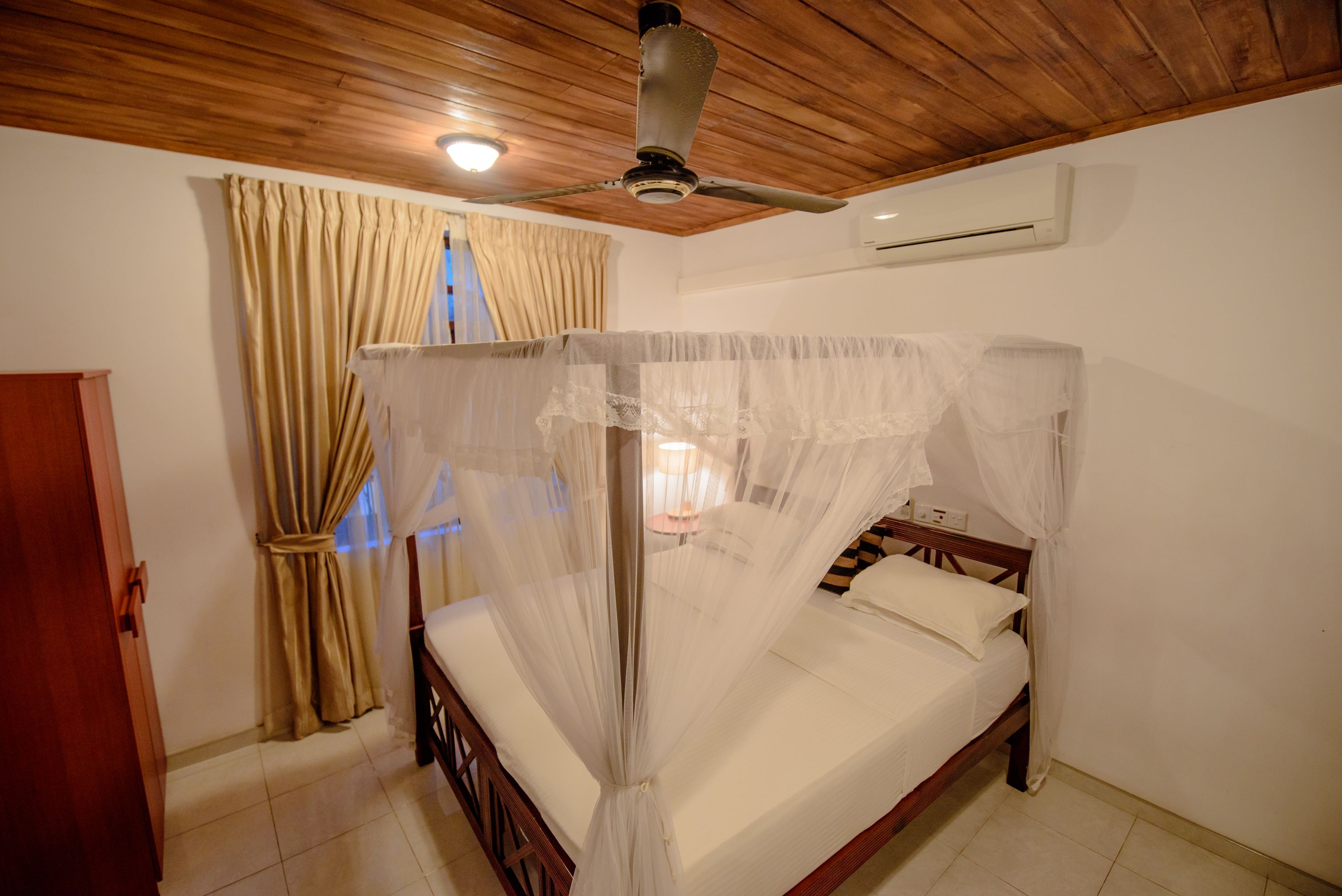 A bedroom with a four-poster bed, wooden ceiling, beige curtains, and an air conditioning unit.