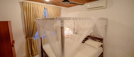 A bedroom with a four-poster bed, wooden ceiling, beige curtains, and an air conditioning unit.