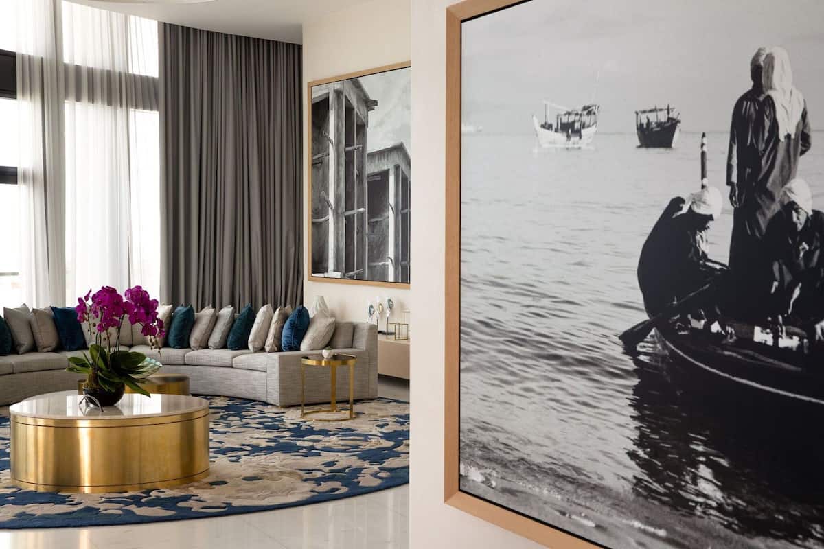 A modern living room with a round gold coffee table, a sofa with patterned cushions, and a large framed black and white photograph of a coastal scene.