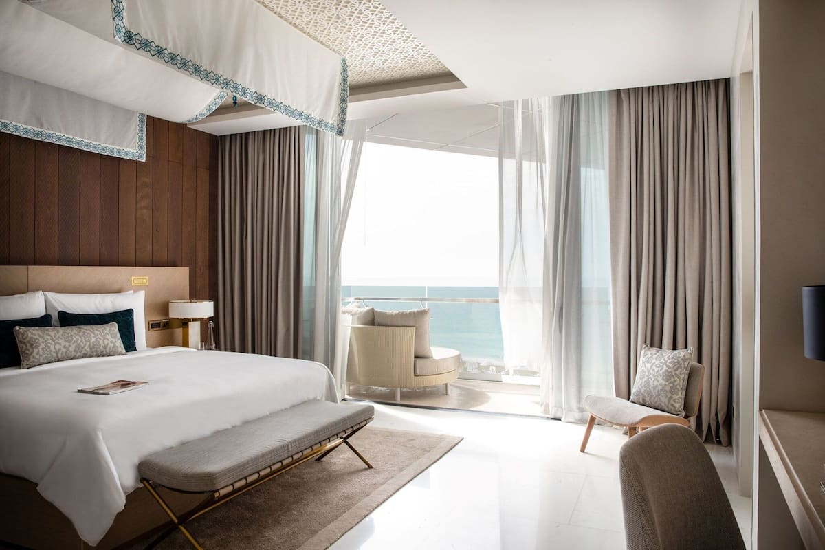A modern hotel room with a large bed, a sitting area with a view of the sea, and a balcony with curtains.