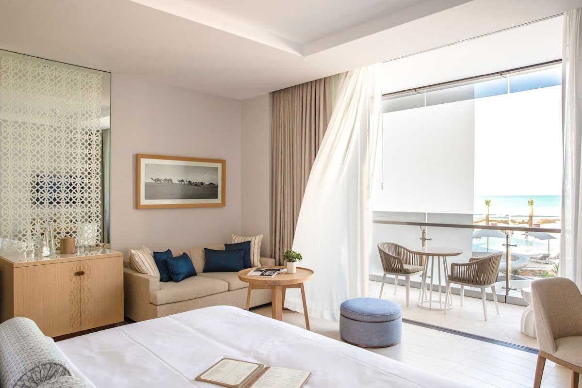 A hotel room with a bed, a sofa, a small table with a plant, a dining area with chairs and a table, and a balcony with a view of the beach.