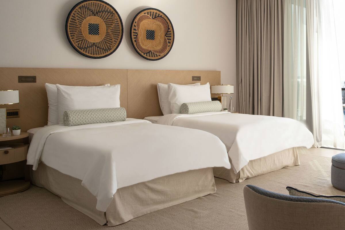 A hotel room with two beds, a nightstand with a lamp, a chair, and two woven wall art pieces.