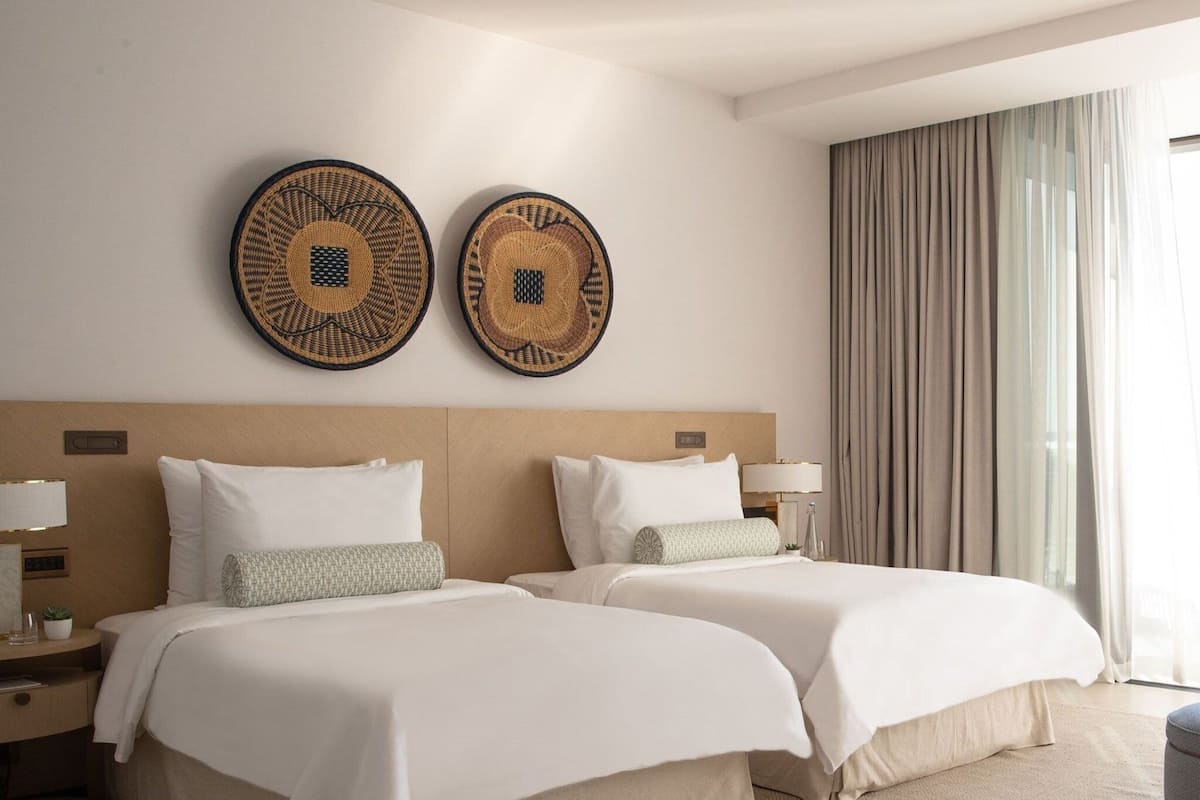 A hotel room with two beds, a wooden headboard, a large window with curtains, and two wall-mounted woven art pieces.