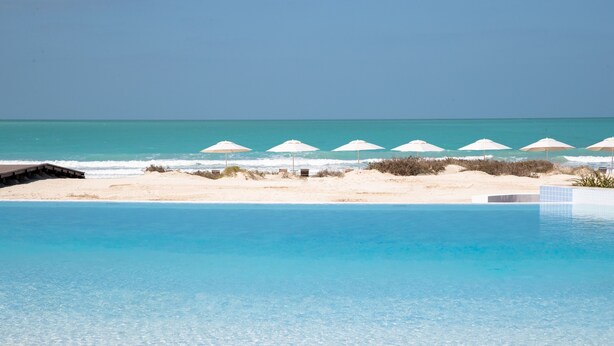 A clear blue pool with a sandy beach and umbrellas in the background. - Jumeirah Saadiyat Island Abu Dhabi (Abu Dhabi)