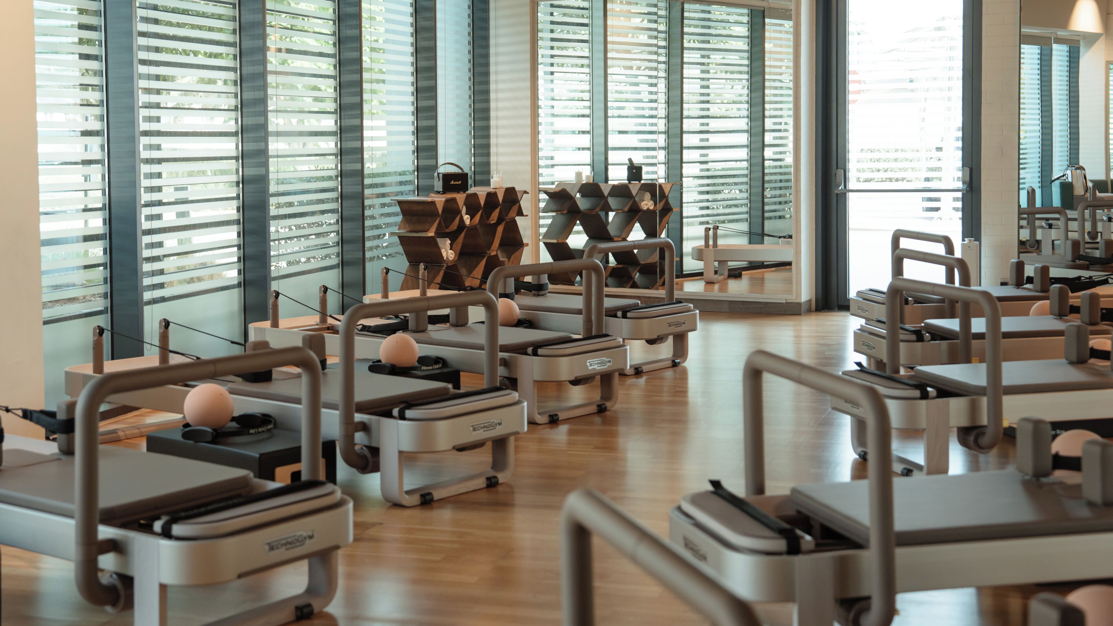 Ein Fitnessstudio mit in Reihen angeordneten Trainingsgeräten, großen Fenstern mit Jalousien und Holzboden.