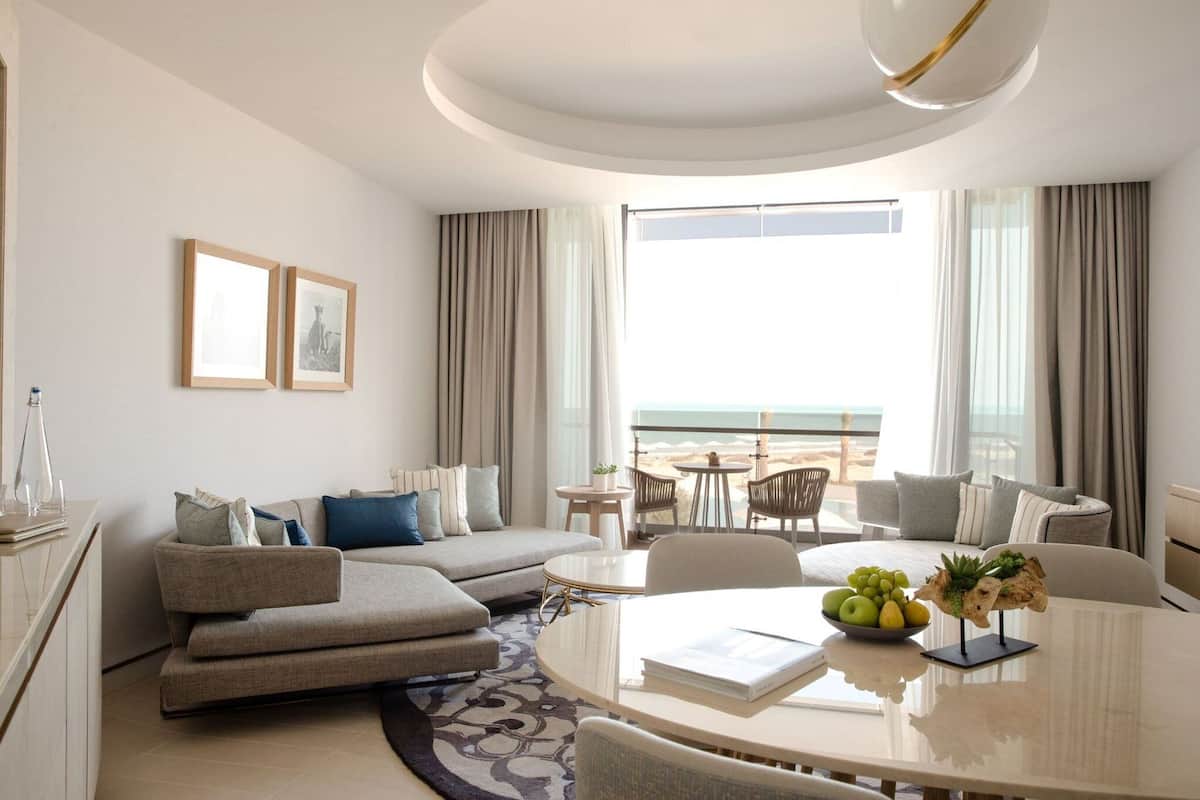 A modern hotel room with a large window offering a view of the beach, a round dining table with a fruit bowl, a sofa, and a small balcony area.