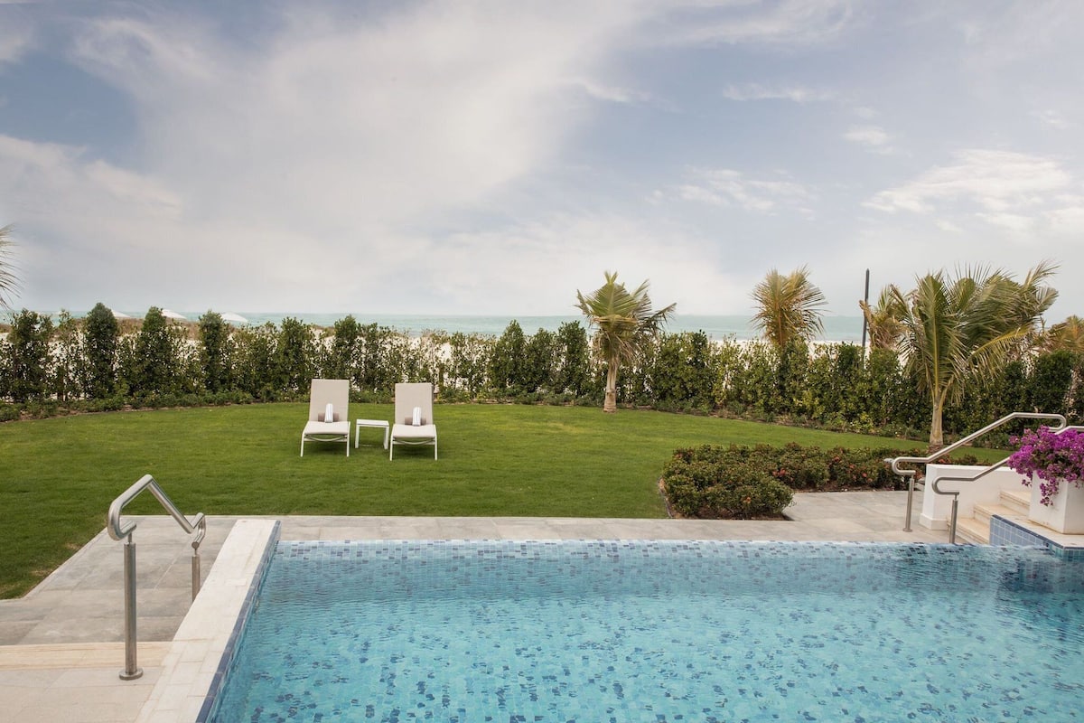A pool area with two lounge chairs, a view of the ocean, and a well-maintained lawn.