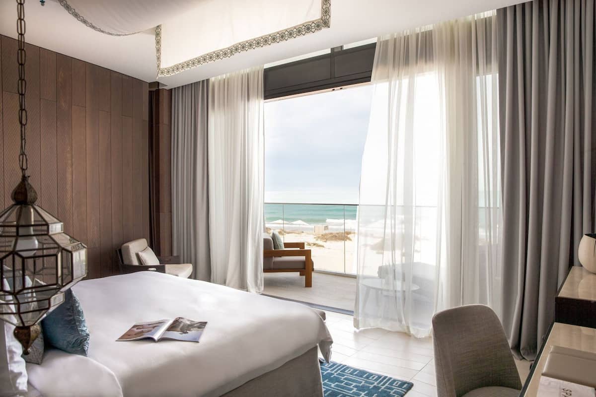 A hotel room with a bed, a chair, a table, and a view of the beach and ocean.