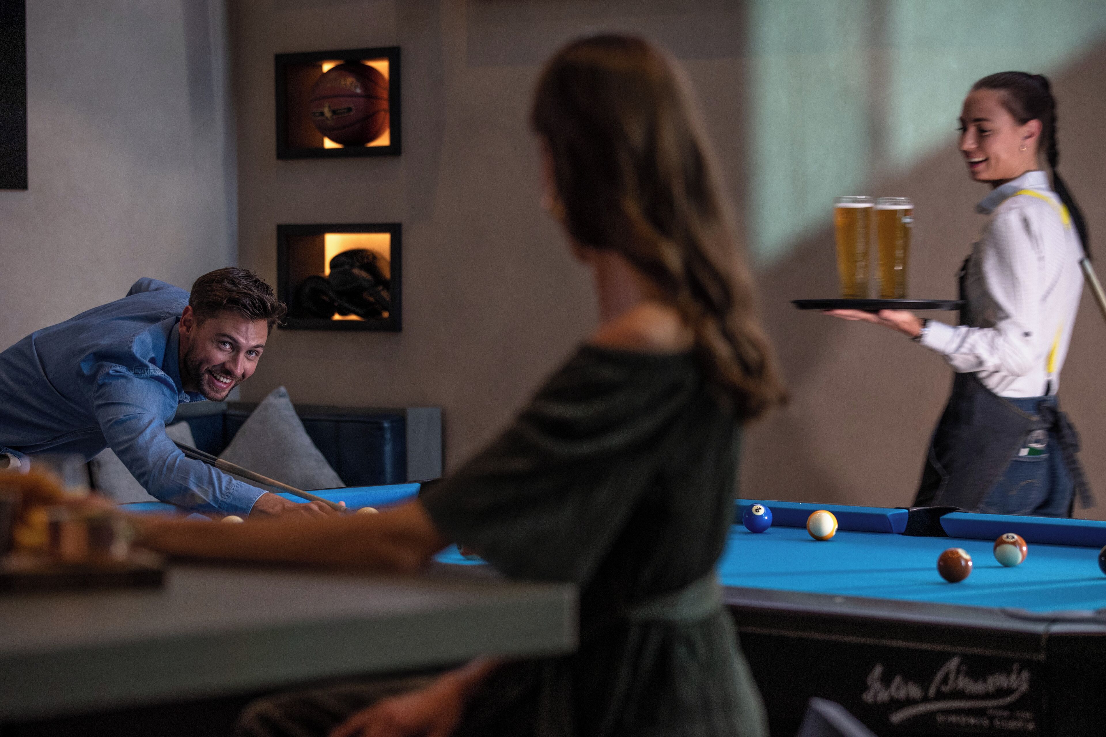 Una mesa de billar con bolas, una persona sirviendo bebidas y cuadros en la pared.