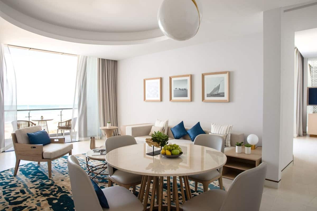 A modern hotel room with a round dining table, comfortable seating, a large window with a view, and a balcony.
