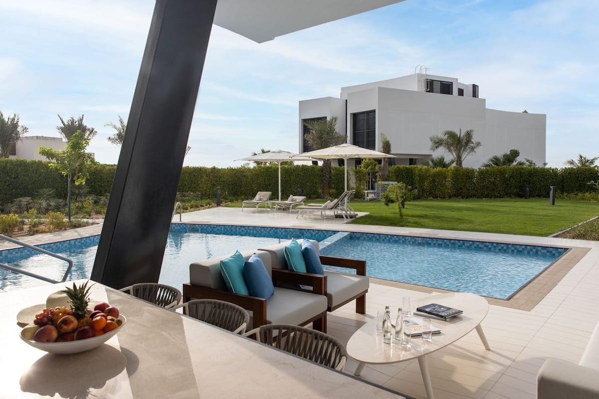 A modern outdoor pool area with lounge chairs and a table, a bowl of fruit, and a white building in the background.