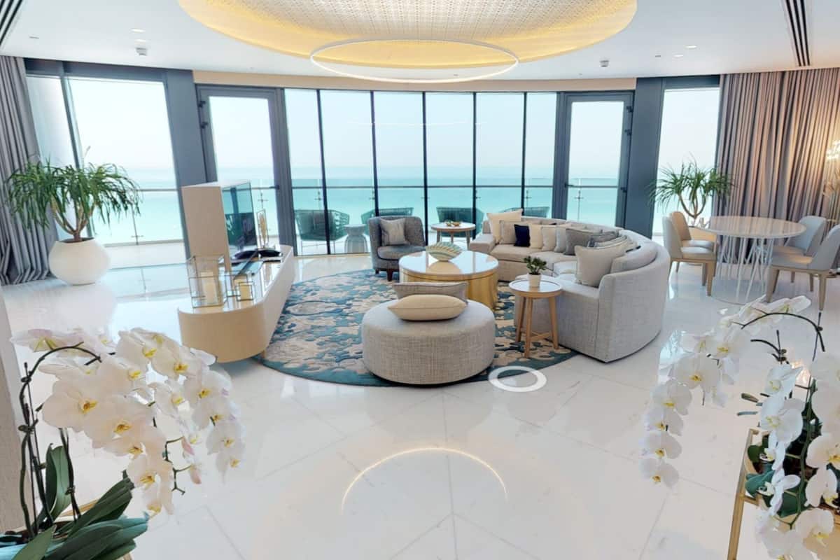 A spacious living room with a large sofa, a coffee table, and a TV. There are potted plants and a view of the ocean through large windows.