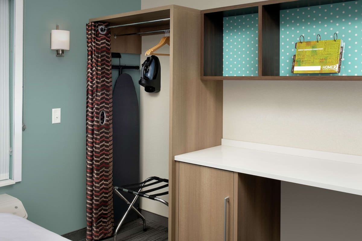 Une chambre d’hôtel de taille réduite, comprenant un lit, un bureau avec une chaise, un porte-manteau et une étagère avec un calendrier.