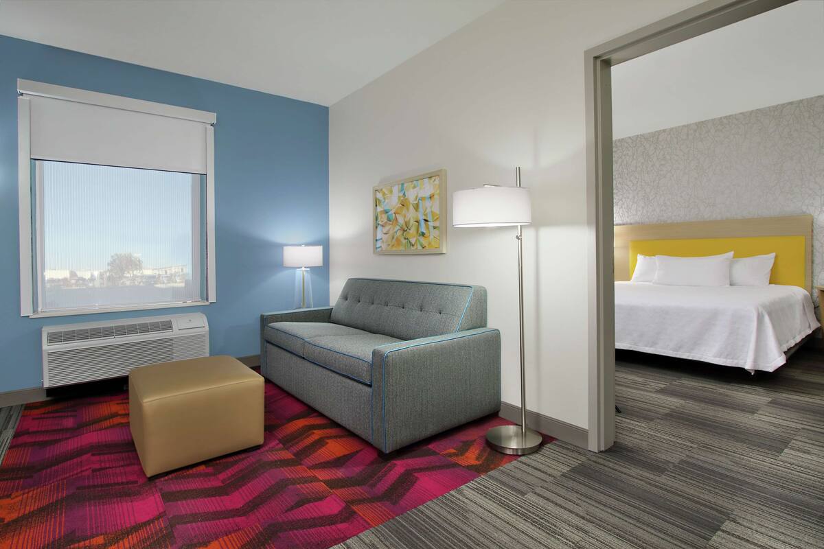 Une chambre d’hôtel équipée d’un canapé, d’un pouf, d’une lampe sur pied et d’un lit avec une tête de lit.