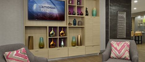 A modern hotel lobby with a flat-screen TV displaying 'National News', a wooden shelving unit with decorative vases, two grey armchairs with patterned cushions, and a 'Home2 Guarantee' sign.