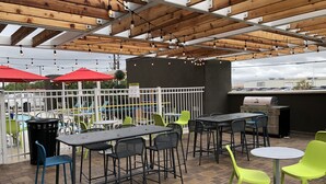 An outdoor patio with tables and chairs, a barbecue grill, and a pool in the background. - Home2 Suites by Hilton Port Arthur, TX (Port Arthur)