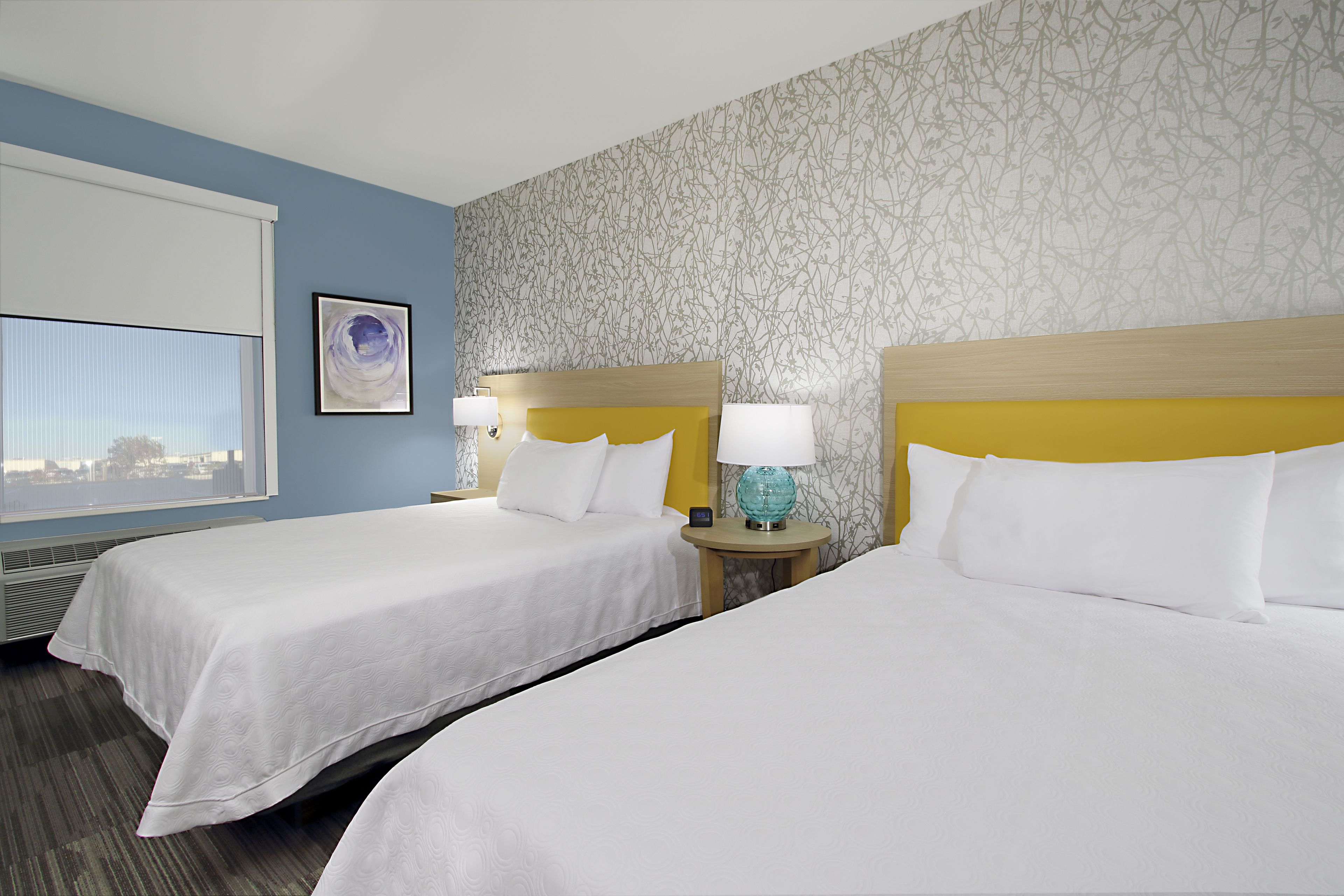 A hotel room with two beds, a window with blinds, a wall with a floral pattern, and a nightstand with a lamp.