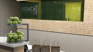 A reception desk with a display of green apples and three brown paper thank you bags. - Home2 Suites by Hilton Port Arthur, TX (Port Arthur)