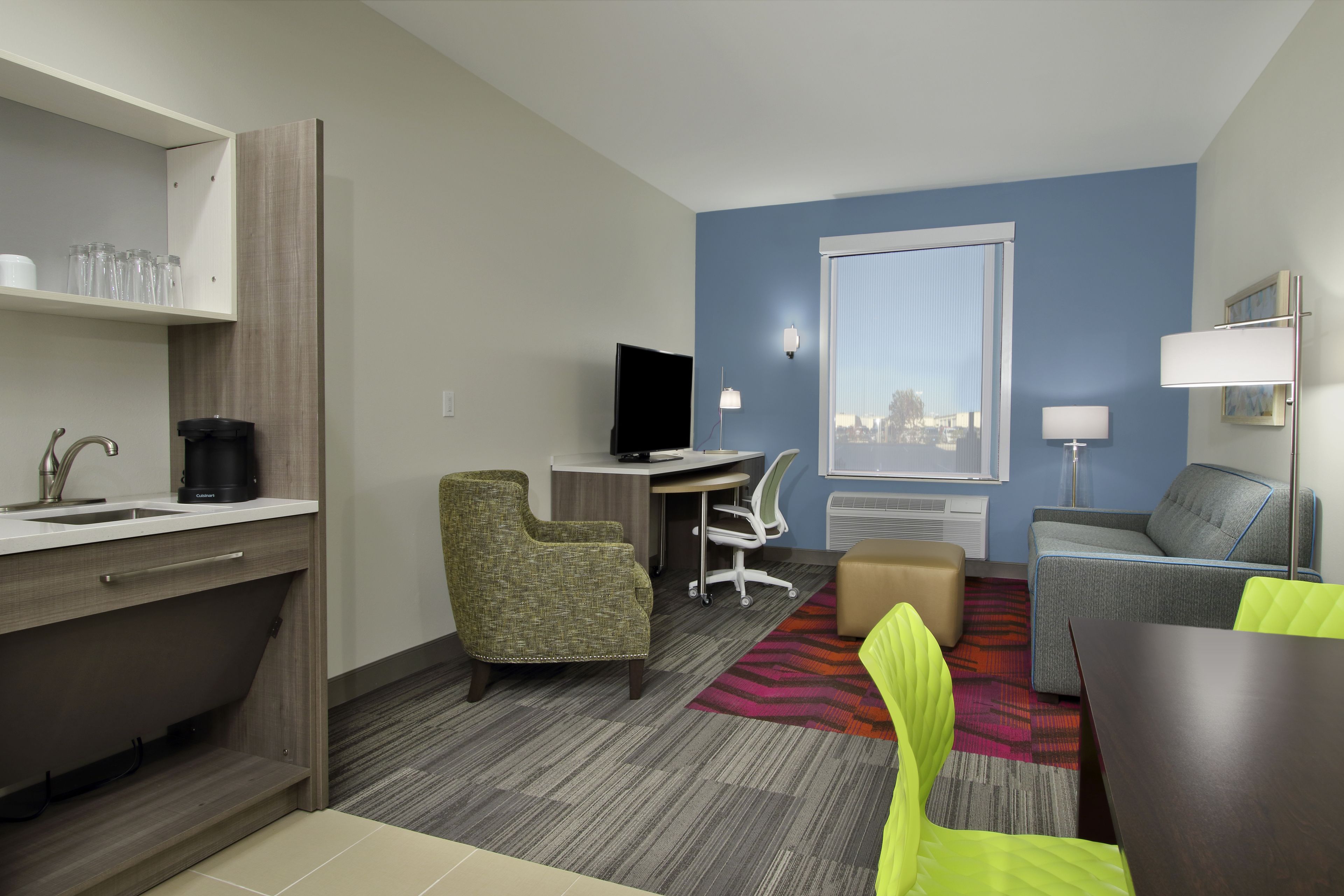 Een hotelkamer met een kitchenette, een bureau, een stoel, een bank en een groot raam.