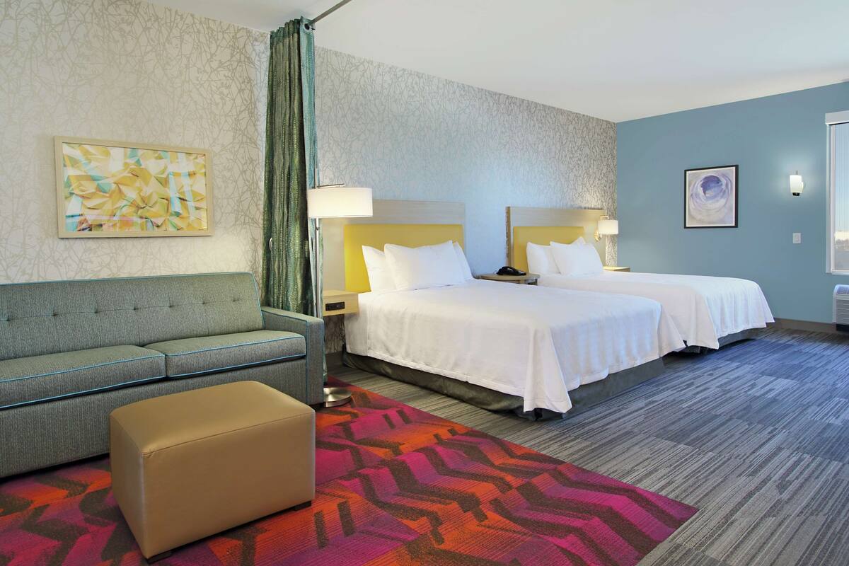 Une chambre d’hôtel avec deux lits, un canapé, un petit pouf et un tapis coloré.