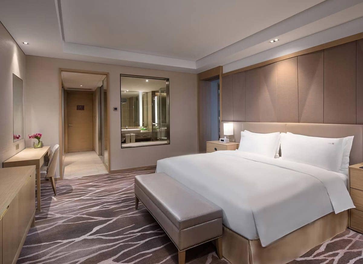 A hotel room with a large bed, a desk, a bench, a mirror, and a bathroom visible through an open door.