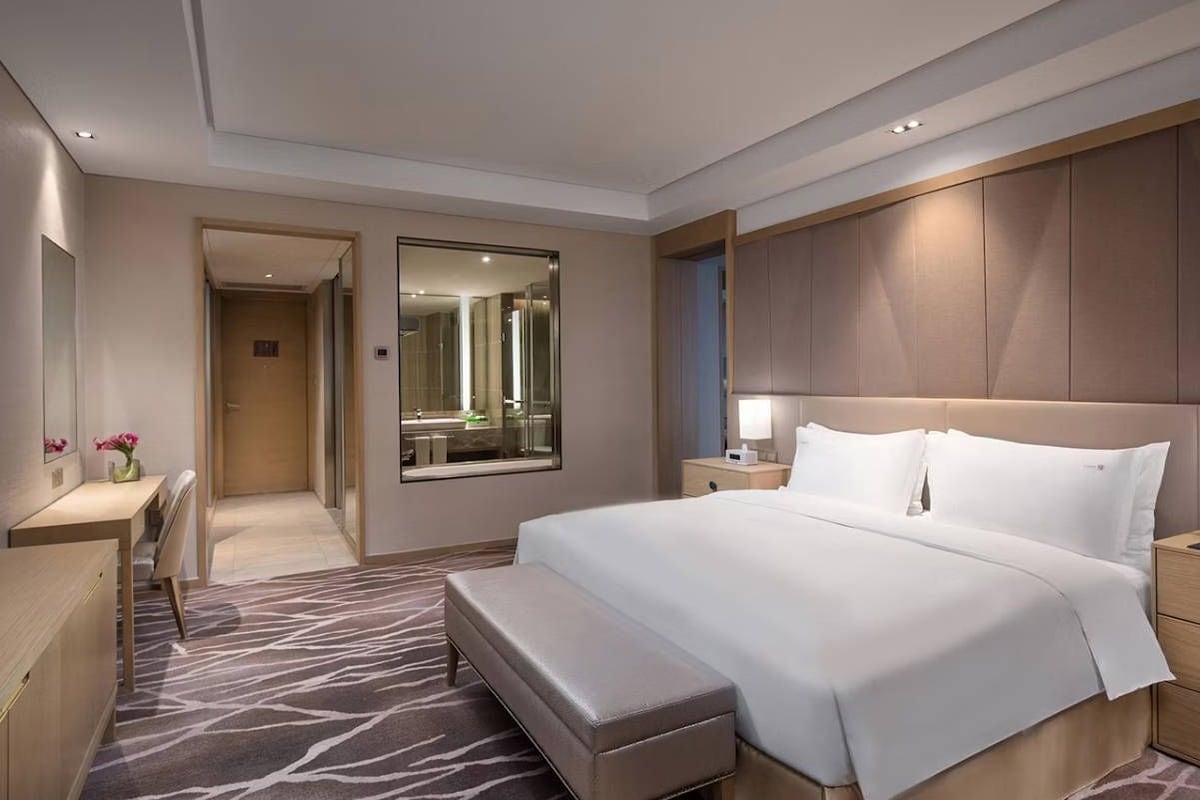 Une chambre d’hôtel avec un grand lit, un bureau, une banquette, un miroir et une salle de bain visible à travers une porte ouverte.
