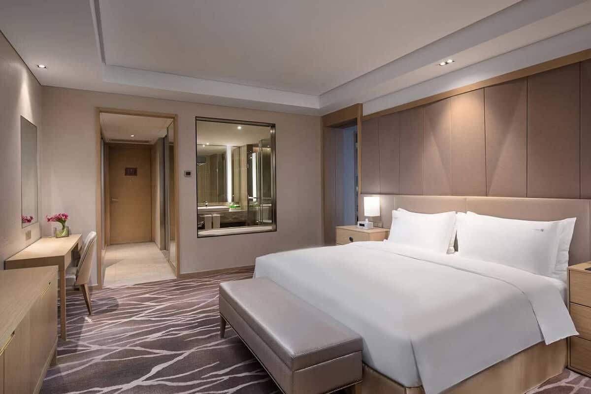 Une chambre d’hôtel avec un grand lit, un bureau, une banquette, un miroir et une salle de bain visible à travers une porte ouverte.