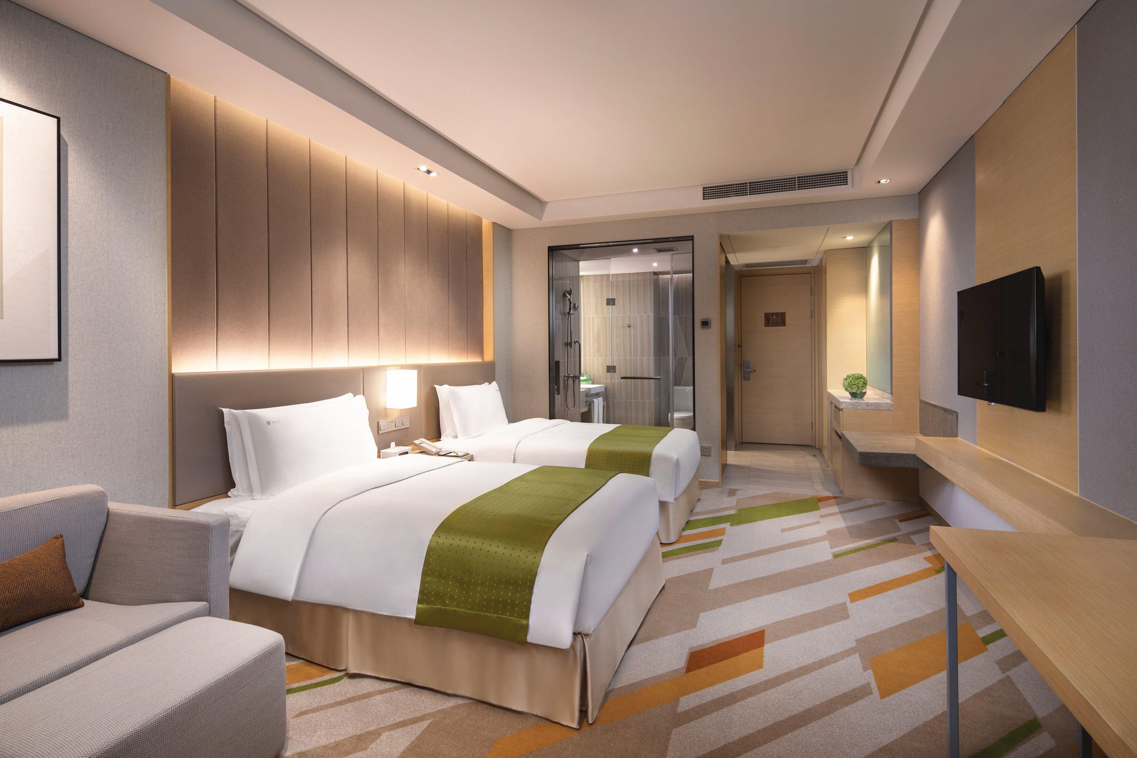 A hotel room with two beds, a sofa, a TV, and a bathroom visible through a glass door.