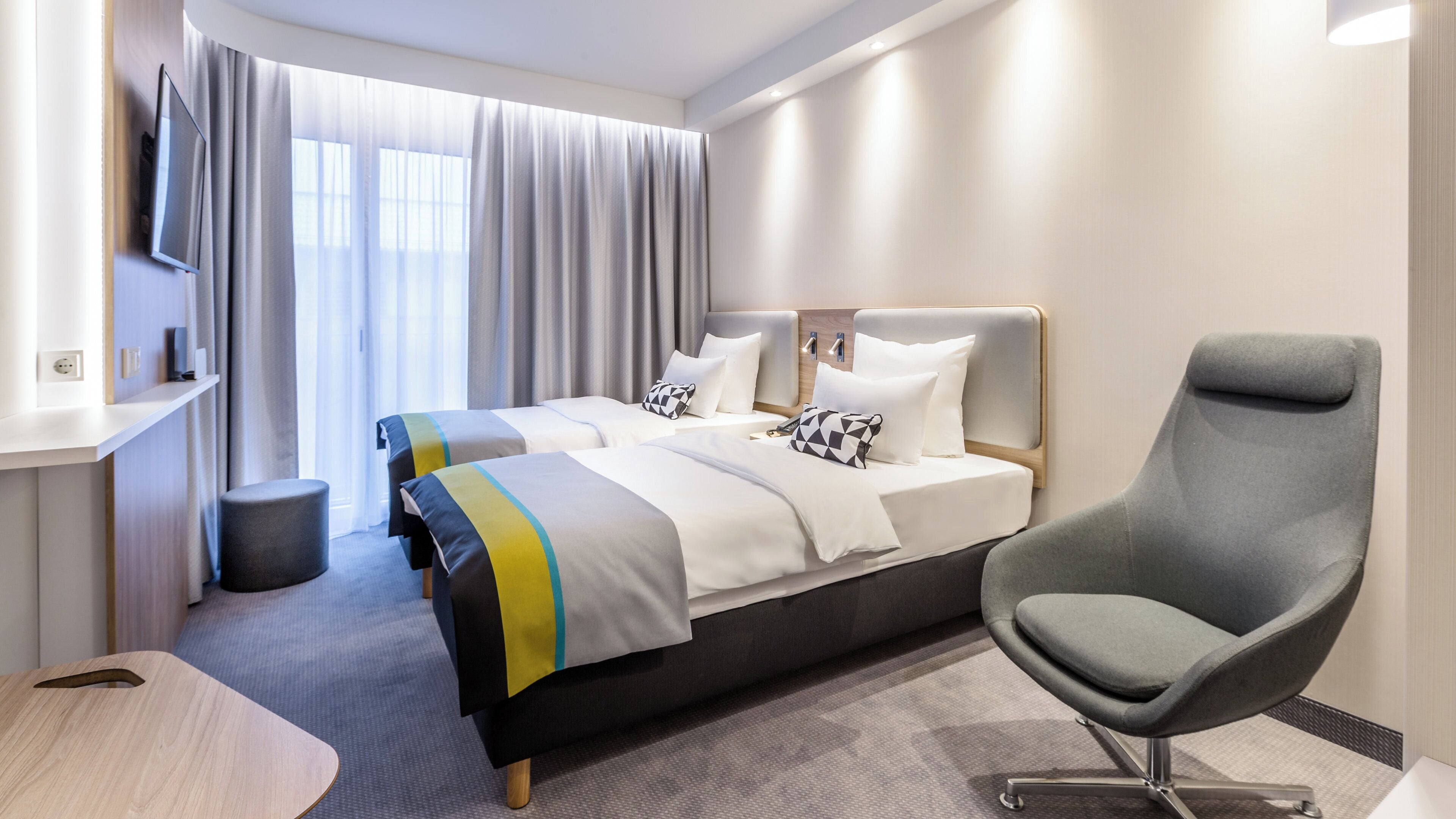 A modern hotel room with two beds, a grey armchair, a TV, and a large window with curtains.