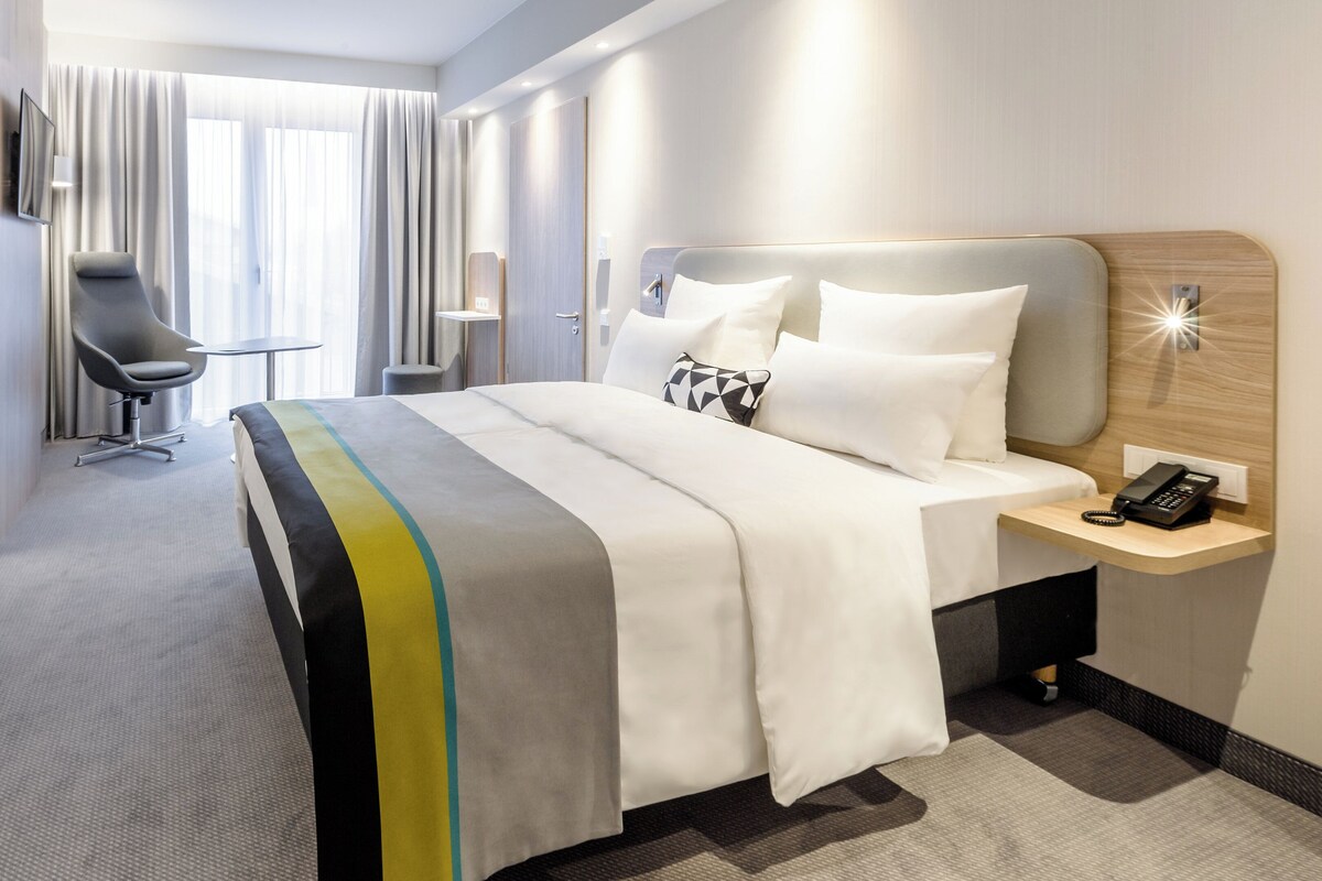 Een hotelkamer met een bed, nachtkastje met telefoon, stoel en bureau.