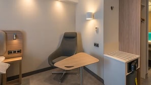 Een moderne hotelkamer met een enkele grijze stoel, een klein houten tafeltje en inbouwkasten met een spiegel en opbergruimte.
