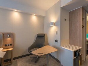 Een moderne hotelkamer met een enkele grijze stoel, een klein houten tafeltje en inbouwkasten met een spiegel en opbergruimte.