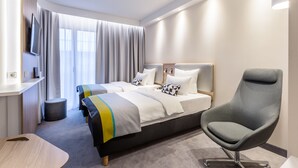 Een moderne hotelkamer met twee bedden, een grijze fauteuil, een televisie aan de muur en een groot raam met gordijnen.
