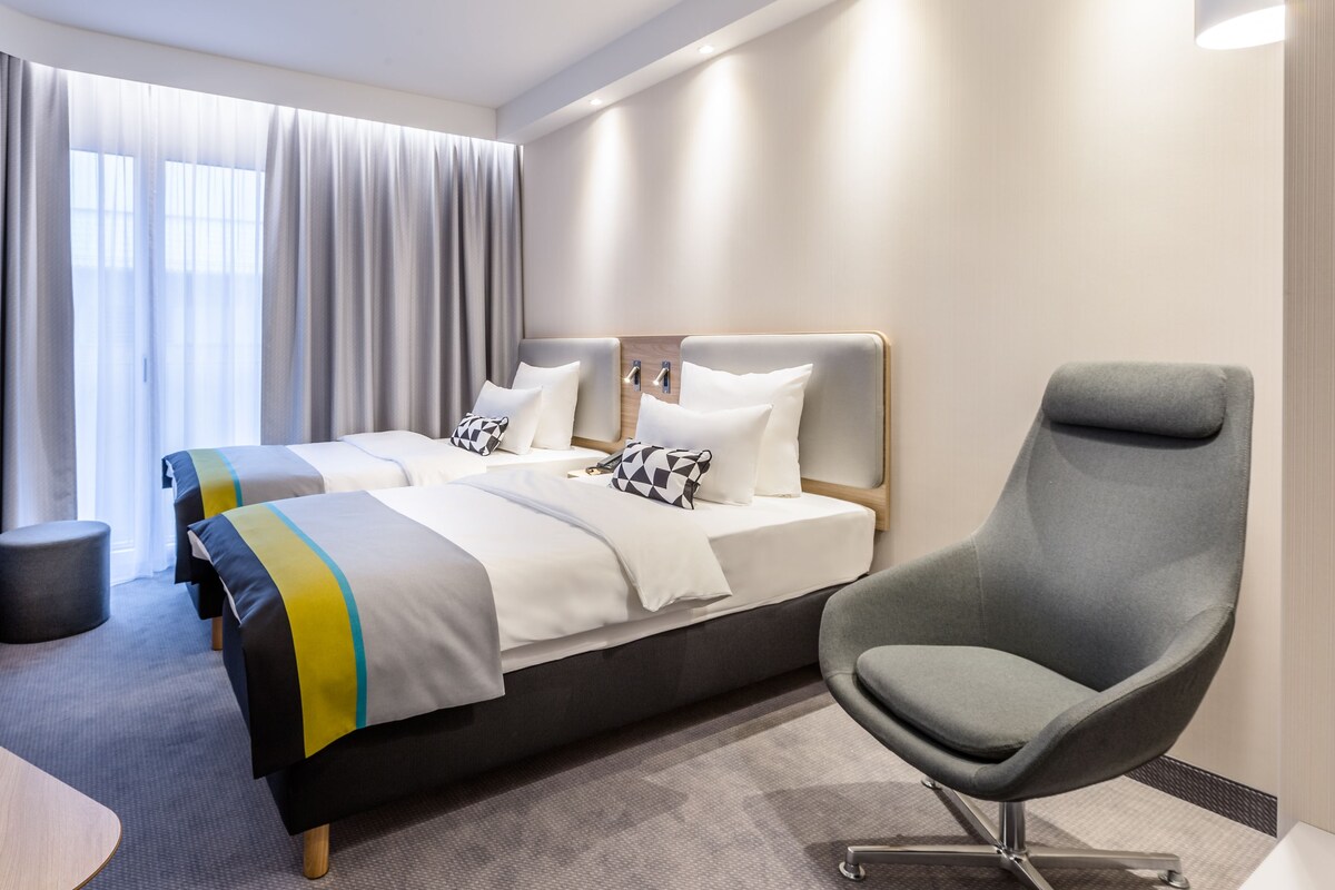 Une chambre d’hôtel moderne avec deux lits, un fauteuil gris, un téléviseur fixé au mur et une grande fenêtre avec des rideaux.