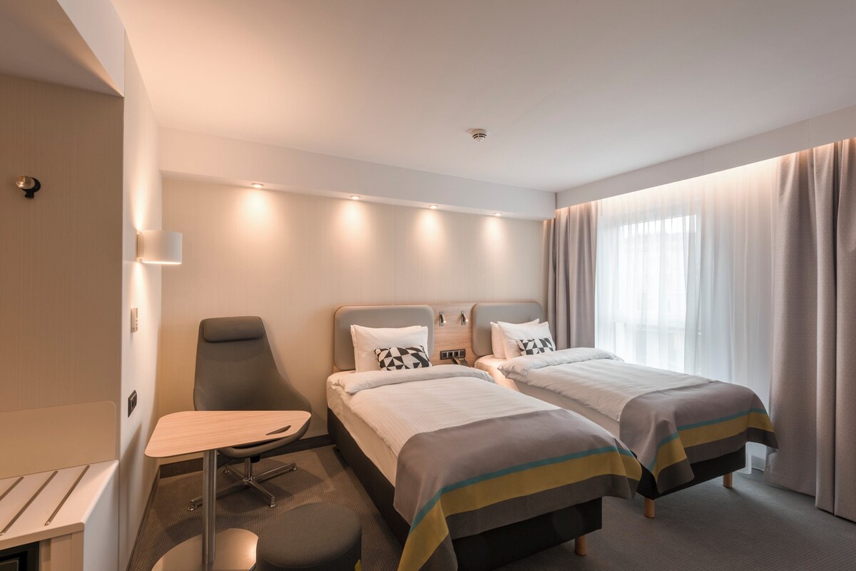 Een hotelkamer met twee bedden, een bureau, een stoel en een raam met gordijnen.
