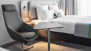 Een moderne hotelkamer met een groot bed, een grijze fauteuil, een klein tafeltje en een nachtkastje met een lamp.