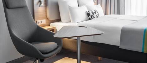 A modern hotel room with a large bed, a grey armchair, a small table, and a nightstand with a lamp.
