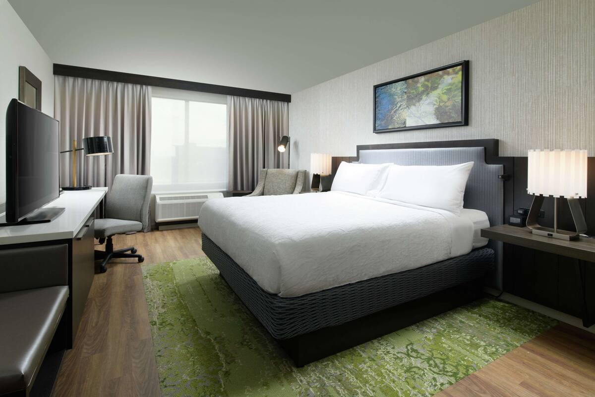 A hotel room with a large bed, a desk with a computer, a chair, a TV, and a window with curtains.